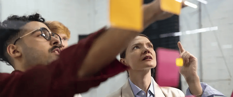 Equipe de profissionais reunida em um ambiente moderno, colando e organizando post-its coloridos em um quadro de vidro transparente, simbolizando trabalho em equipe, planejamento e inovação.